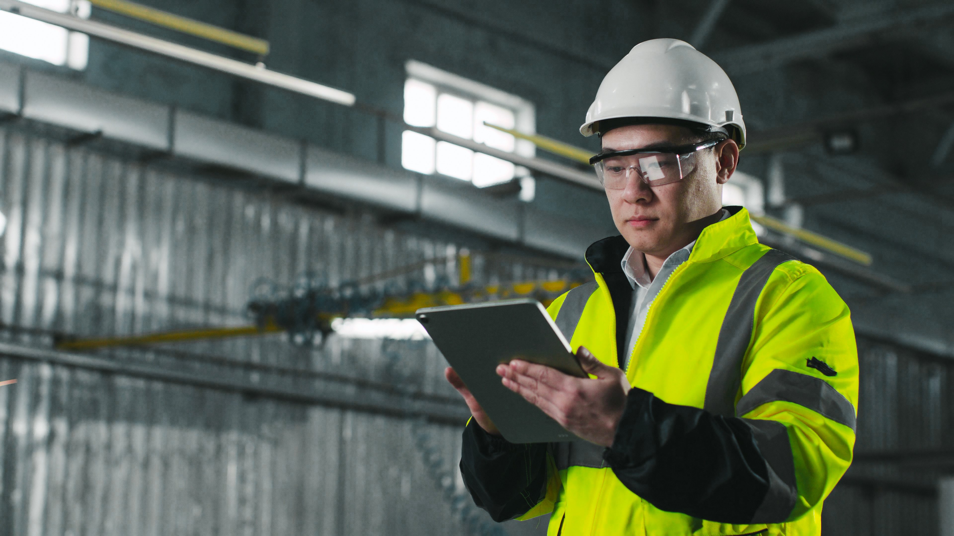 Asian engineer looks at screen of tablet. Worker wearing yellow vest, glasses and hard hat.Male stands on background of workflow. Man at workplace.