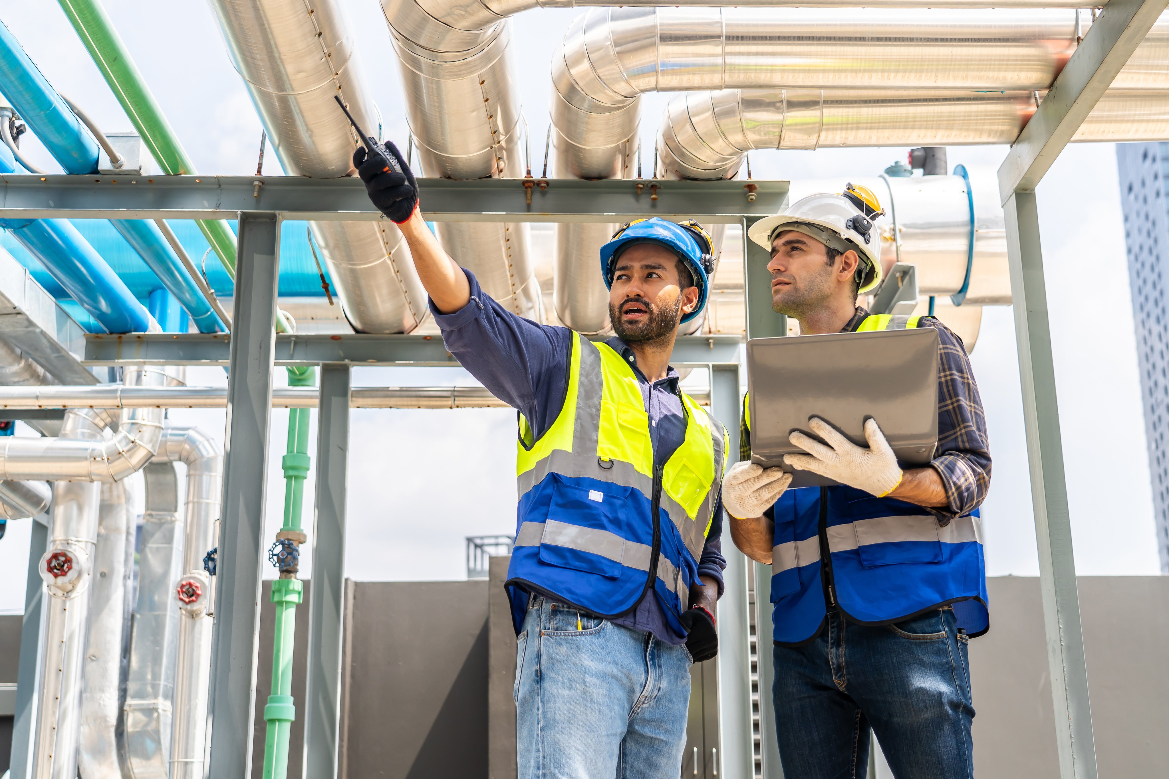 Collaboration, Two young construction workers discuss plans and use laptop computer while reviewing blueprints on a rooftop. They wear safety gear and oversee intricate piping systems in factory