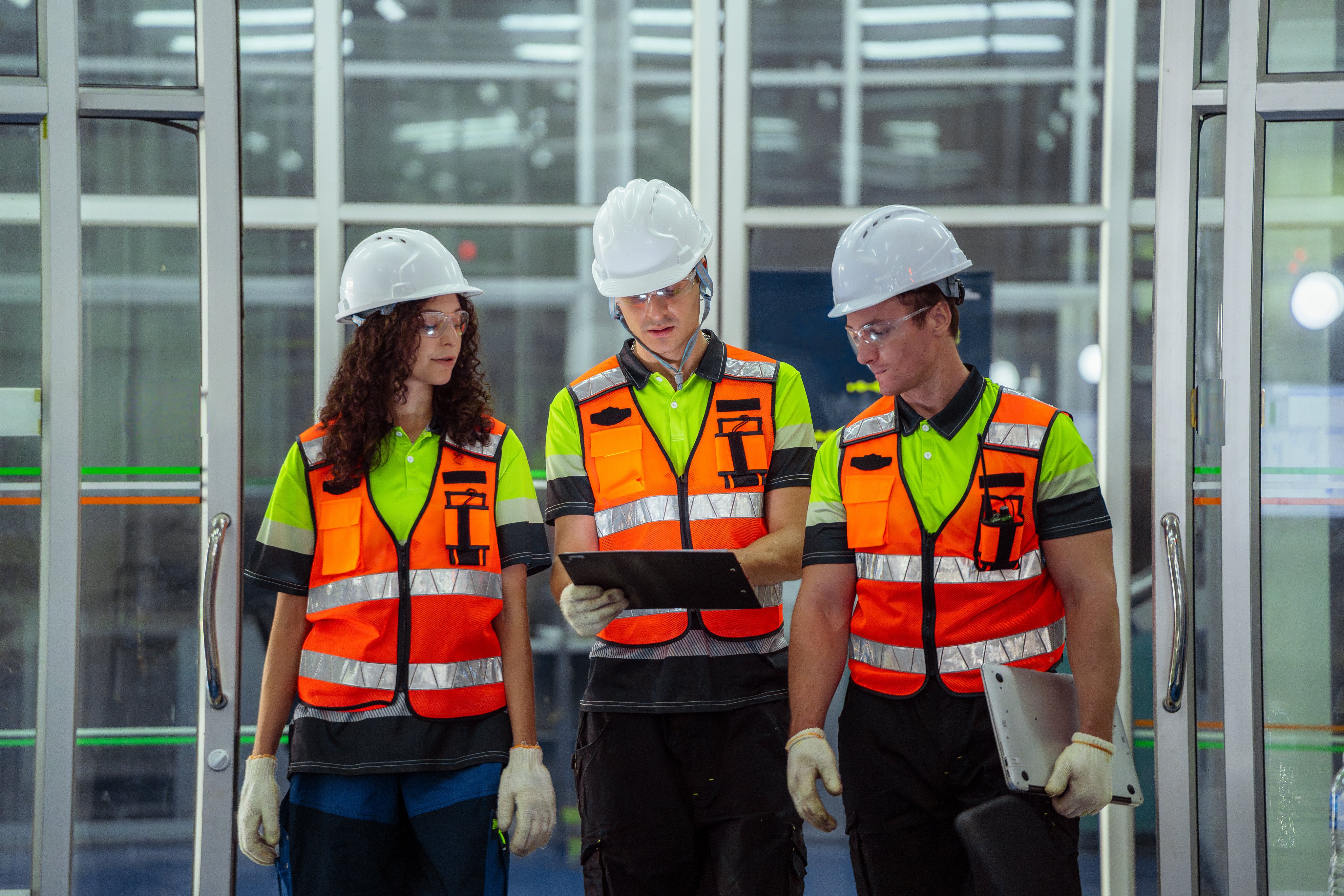 Three people wearing safety vests and hard hats. One of them is holding a laptop. They are standing in front of a door
