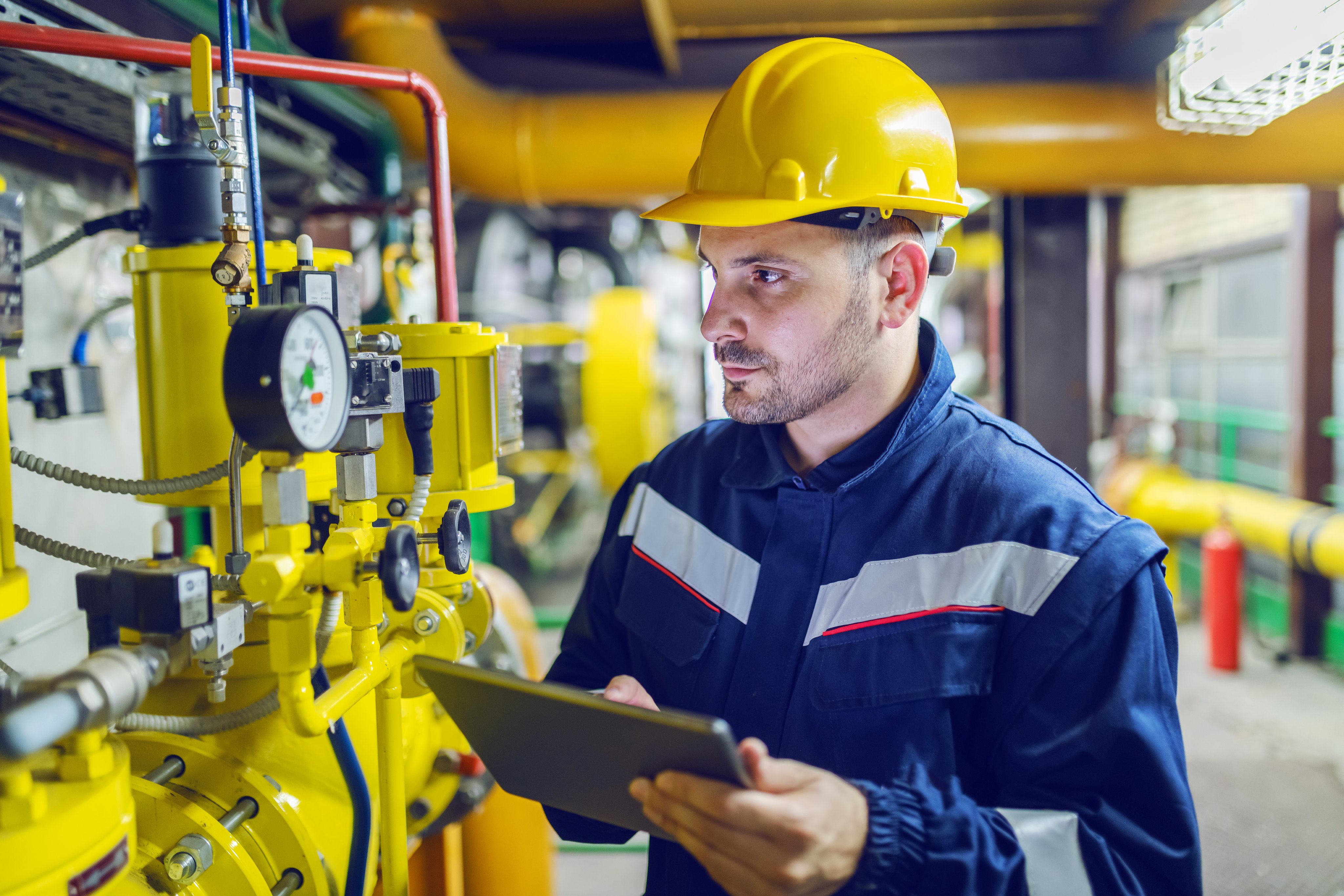 Serious caucasian unshaven worker in protective uniform and with hardhat using tablet for checking temperature in pipes. Factory interior.