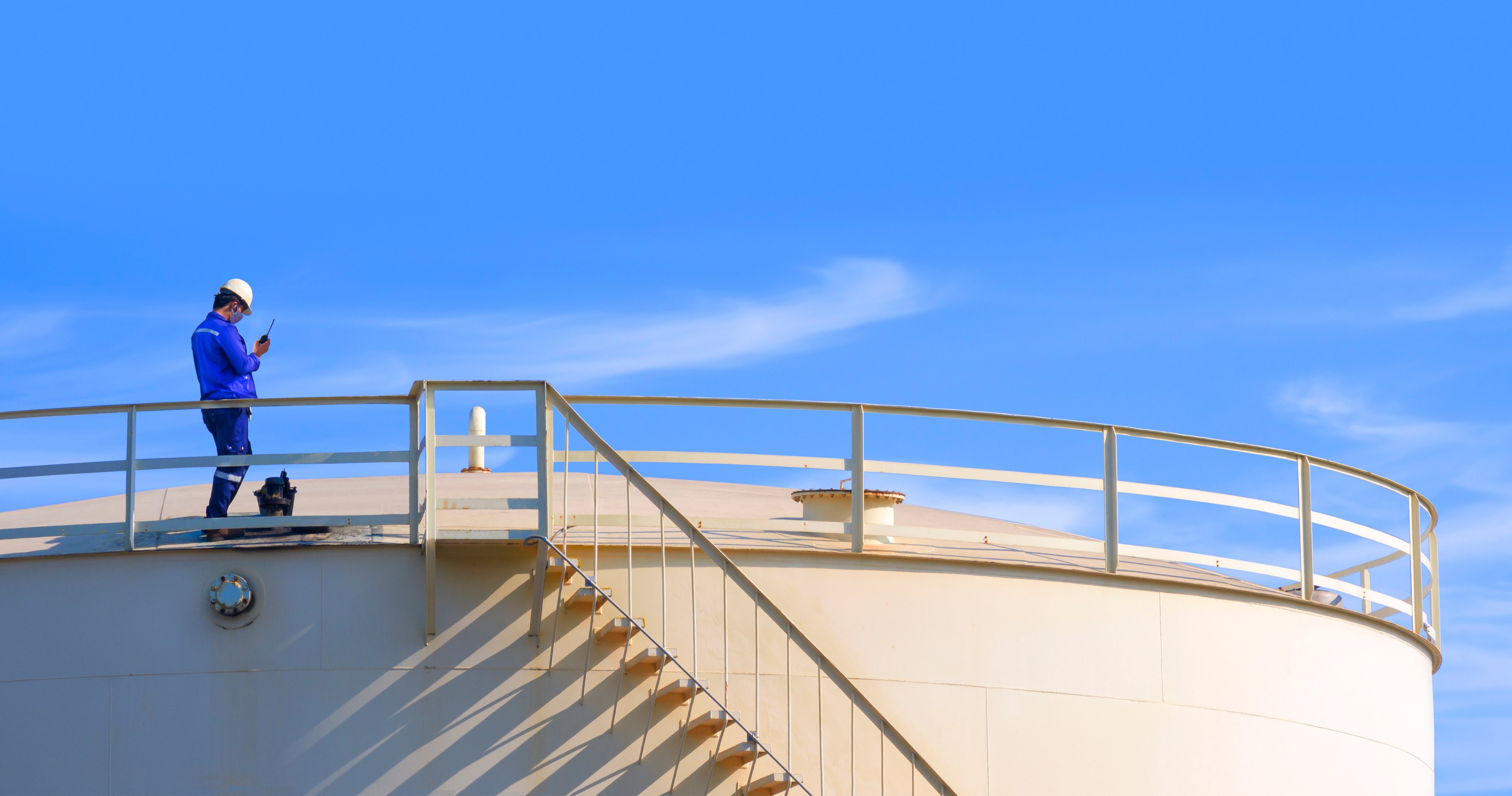 Engineer and worker are working to check oil quality on top of big fuel storage tank against blue clear sky background