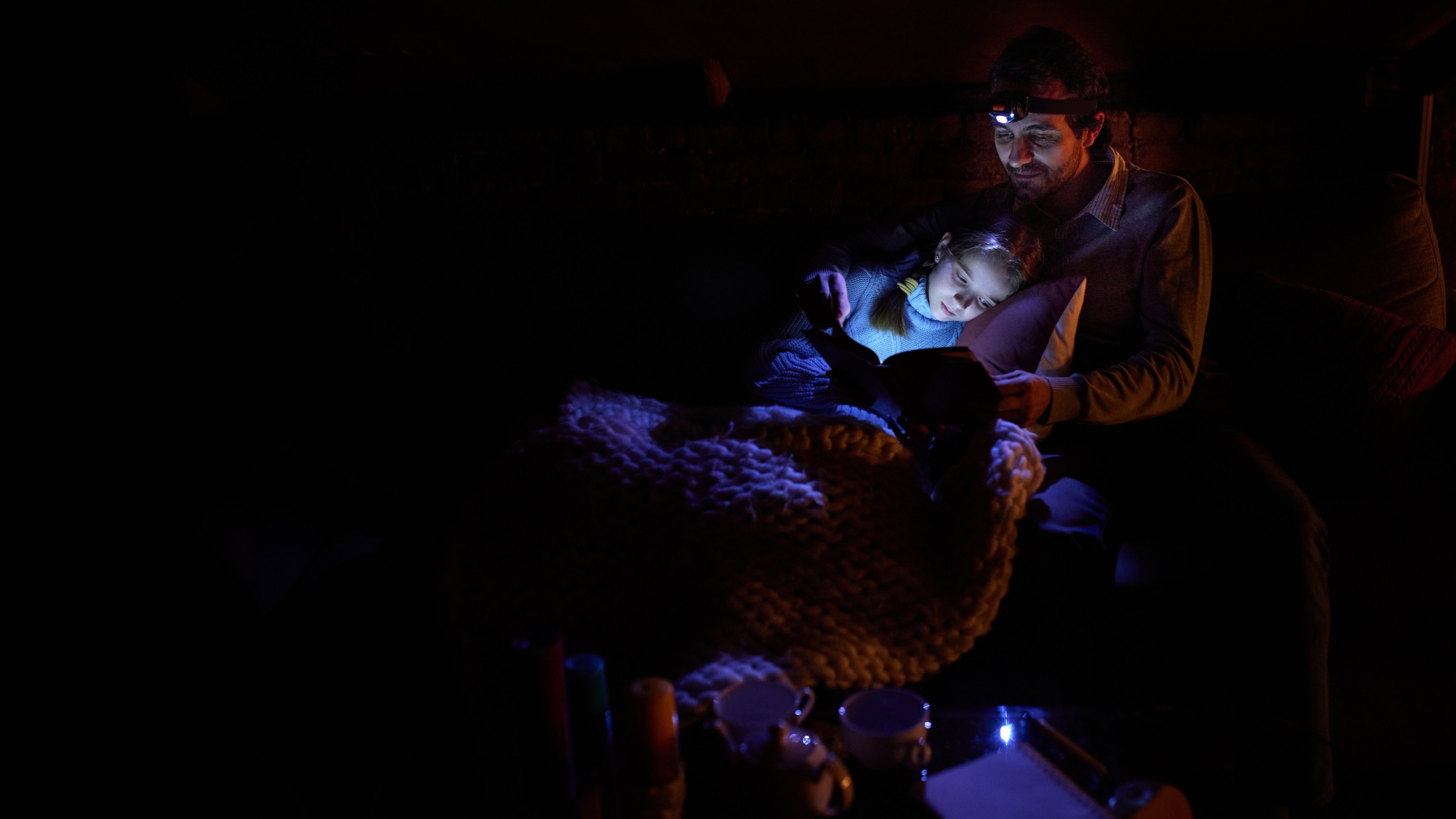 Father with his little daughter sitting on sofa at home in the evening and reading book with forehead battery lamp. No electricity, blackout. Concept of power outage. Banner. Copy space for ad, text