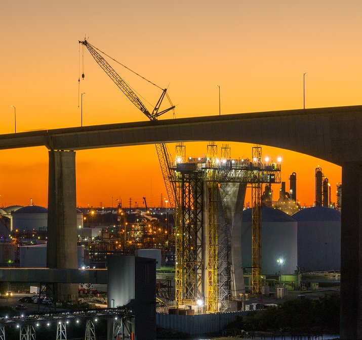Overpass bridge construction underway at sunset with crane in operation and oil refineries in the background.